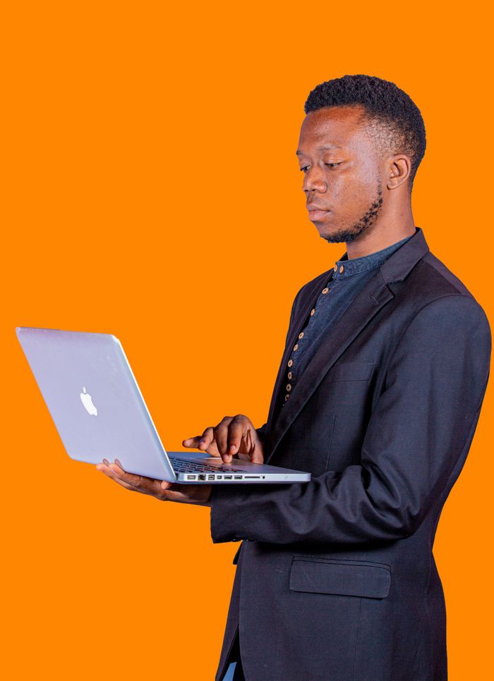 Services Man in black suit using a laptop against an orange studio backdrop, displaying focus and engagement.