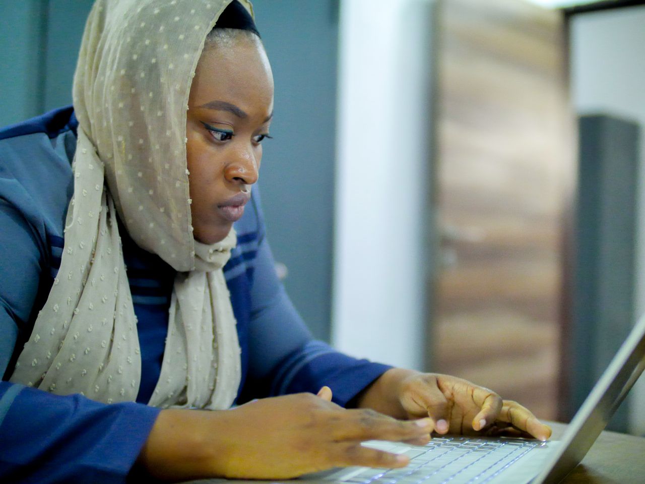 Services Woman in headscarf intensely working on a laptop indoors, demonstrating focus and determination.